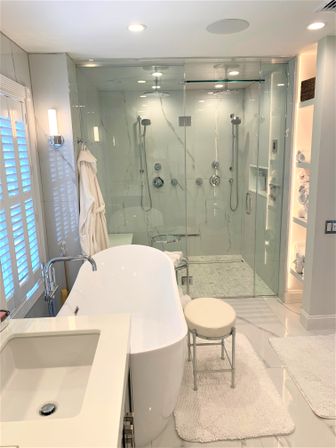 Spa-like modern master bathroom featuring a white freestanding tub, glass-enclosed dual rain shower with marble-look tile, vanity sink, plush rugs and stacked towels.