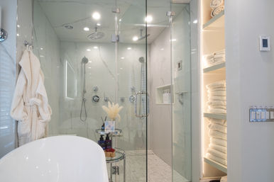 Luxury spa-like bathroom with glass-enclosed marble shower (rain and handheld heads), freestanding tub, plush robe and neatly stacked towels on lit shelves.