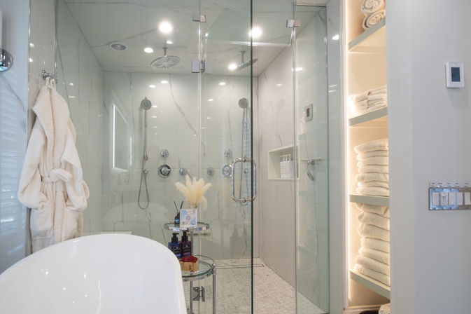 Luxury spa-like bathroom with glass-enclosed marble shower (rain and handheld heads), freestanding tub, plush robe and neatly stacked towels on lit shelves.