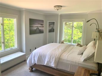 Bright, serene master bedroom with white linens on a wooden platform bed, large windows framing lush green trees, tufted window bench, lakeside pier wall art, and adjustable reading lamps