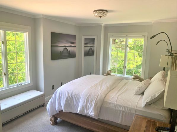 Bright, serene master bedroom with white linens on a wooden platform bed, large windows framing lush green trees, tufted window bench, lakeside pier wall art, and adjustable reading lamps
