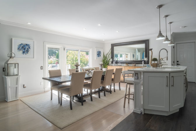 Bright open-concept dining room and kitchen with a long dark wood table seating eight on beige upholstered chairs, large light rug, white island with pendant lights and a stainless gooseneck faucet, oversized mirror and sliding glass doors opening to a sunlit garden.