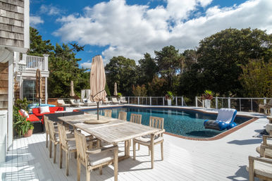 Sunny coastal-style backyard pool deck with weathered wood dining table and umbrella, lounge chairs, red outdoor sofa and inflatable pool float beside a blue pool surrounded by trees.