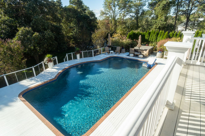 Aerial view of a backyard swimming pool with sparkling blue water, white wooden deck and brick edging, patio seating with umbrellas, an inflatable lounger, and lush trees and shrubs surrounding the yard.