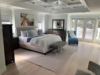Sunlit master bedroom with white bedding and upholstered headboard, blue-striped pillows, tufted teal chaise, French doors to balcony, light hardwood floors and crystal chandelier.