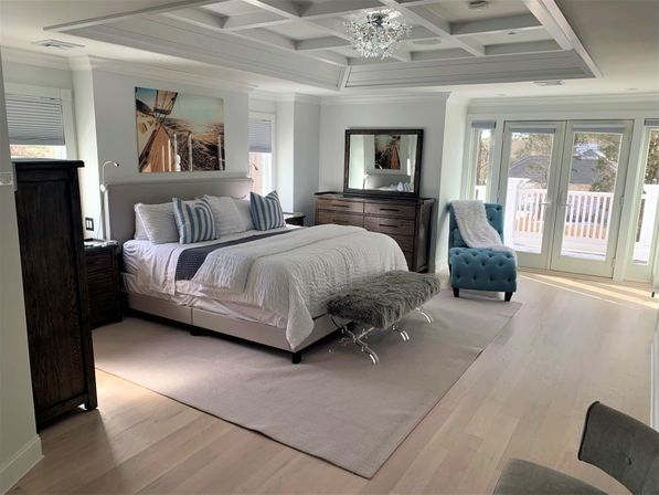 Sunlit master bedroom with white bedding and upholstered headboard, blue-striped pillows, tufted teal chaise, French doors to balcony, light hardwood floors and crystal chandelier.