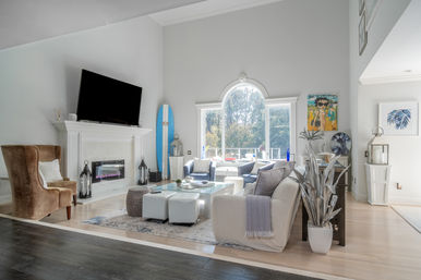 Bright modern coastal living room with vaulted ceiling, arched window, TV above white fireplace, blue surfboard leaning by mantel, neutral seating, glass coffee table and light wood floors.