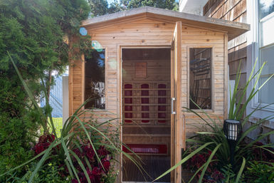 Cozy wooden backyard sauna cabin with open door revealing red infrared panels and a wooden bench, nestled among lush garden plants, tall shrubs and a decorative lantern.