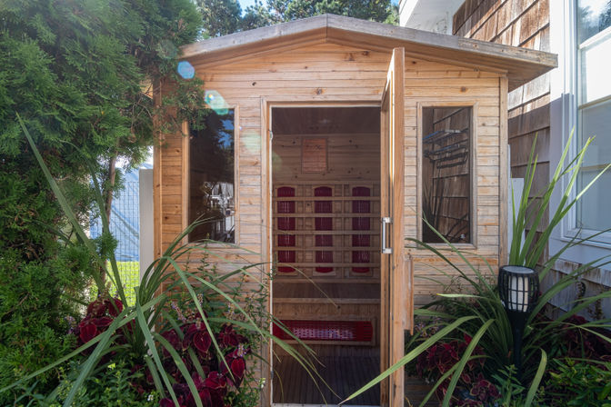 Cozy wooden backyard sauna cabin with open door revealing red infrared panels and a wooden bench, nestled among lush garden plants, tall shrubs and a decorative lantern.