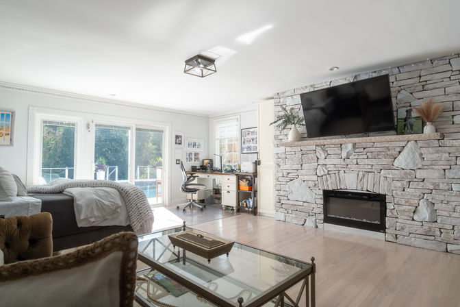 Bright modern living room-bedroom with stacked stone fireplace and wall-mounted TV, glass coffee table, bed and home office by sliding doors to a backyard pool
