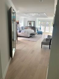 Sunlit modern master bedroom with light hardwood floors, white bedding and striped pillows, tufted turquoise chaise by glass balcony doors, wooden dresser with mirror, coffered ceiling and small crystal chandelier.