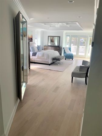 Sunlit modern master bedroom with light hardwood floors, white bedding and striped pillows, tufted turquoise chaise by glass balcony doors, wooden dresser with mirror, coffered ceiling and small crystal chandelier.