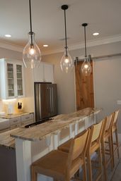 Bright modern farmhouse kitchen with marble-topped island breakfast bar, four woven wooden bar stools, teardrop glass pendant lights, stainless steel refrigerator and sliding barn door.