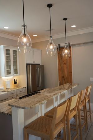 Bright modern farmhouse kitchen with marble-topped island breakfast bar, four woven wooden bar stools, teardrop glass pendant lights, stainless steel refrigerator and sliding barn door.