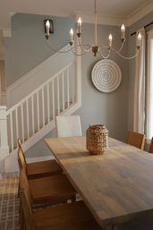 Modern farmhouse dining room with a long wooden table, wicker centerpiece, wooden chairs, white staircase banister, round wall decor and a candle-style chandelier against soft gray walls.