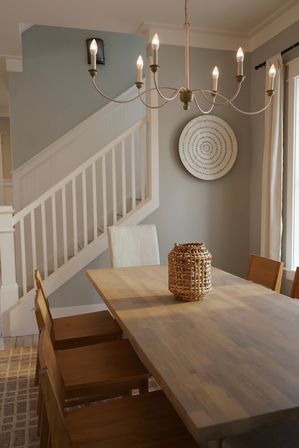 Modern farmhouse dining room with a long wooden table, wicker centerpiece, wooden chairs, white staircase banister, round wall decor and a candle-style chandelier against soft gray walls.