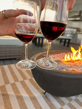 Two red wine glasses clinking above a glowing outdoor fire pit on a striped patio table, decorative stem charms visible — cozy patio evening scene