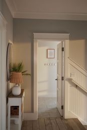 Cozy sunlit entryway and foyer with white paneled staircase, open door to tiled mudroom, wood-top console table, wicker lamp, round mirror and potted greenery.