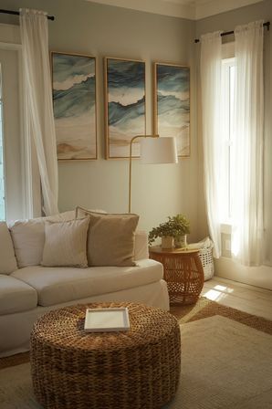 Sunlit coastal‑inspired living room with a white sofa and beige pillows, woven rattan ottoman coffee table, round rattan side table with potted plant, gold floor lamp, sheer curtains, and three ocean‑themed framed prints.
