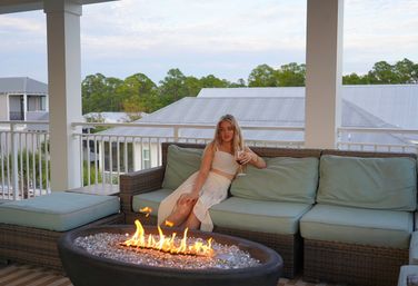 Woman relaxing on a covered outdoor balcony patio, seated on a wicker sofa with pale blue cushions sipping a glass of wine while a modern glass-bead fire pit blazes in the foreground and tree-lined rooftops stretch beyond.