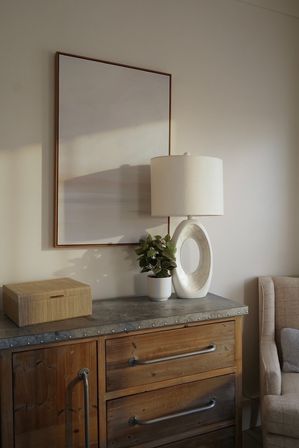 Cozy modern farmhouse bedroom vignette: wooden dresser with metal top, woven storage box, small potted plant, sculptural oval ceramic table lamp and framed minimalist artwork on a neutral wall.