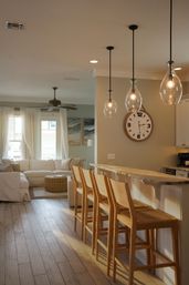 Bright open-plan coastal-style living area and kitchen island with four wooden bar stools, wood countertop, three teardrop glass pendant lights, large round wall clock, neutral sectional sofa and wicker ottoman