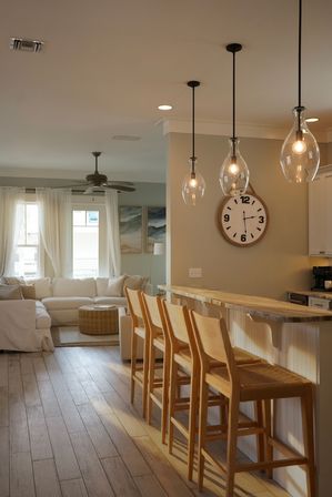 Bright open-plan coastal-style living area and kitchen island with four wooden bar stools, wood countertop, three teardrop glass pendant lights, large round wall clock, neutral sectional sofa and wicker ottoman