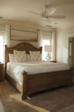 Cozy sunlit master bedroom with wooden sleigh bed, crisp white linens and patterned pillows, ceiling fan, bedside lamp and sheer curtains