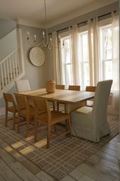 Sunlit dining room with rectangular wood table, mixed wooden and upholstered chairs, woven basket centerpiece, linen curtains, chandelier and neutral coastal-inspired decor.
