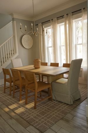 Sunlit dining room with rectangular wood table, mixed wooden and upholstered chairs, woven basket centerpiece, linen curtains, chandelier and neutral coastal-inspired decor.