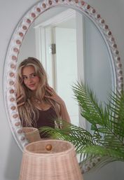 Smiling woman admiring her hair in an oval wall mirror in a bright, coastal-style bedroom with a wicker lamp and green palm plant