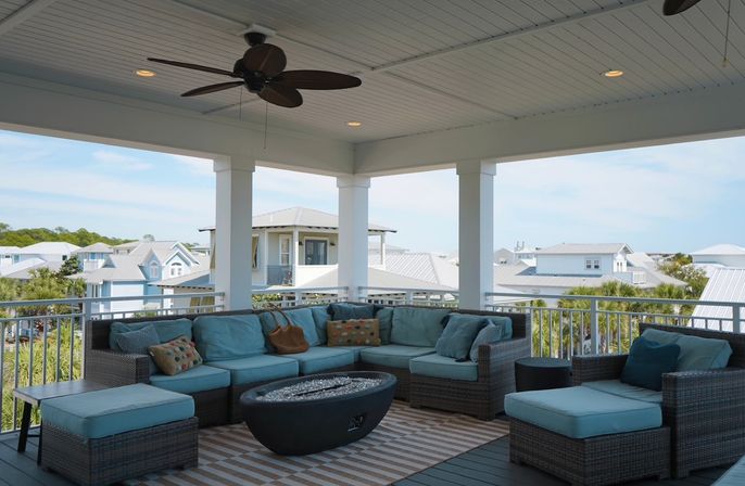Shaded coastal balcony with wicker sectional and turquoise cushions, glass fire pit table, ceiling fan and railing overlooking beach-style houses and palm trees.