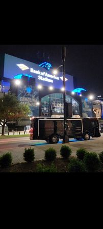 Night shot of a blue-lit football stadium facade in Charlotte, NC with team banners on the exterior and a black shuttle bus parked curbside.