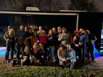 Nighttime outdoor group photo of about 20 adults posing on a grassy lawn in front of a black party/tour bus with blue underglow lighting, wearing casual outfits and hats and holding drinks, smiling and socializing.
