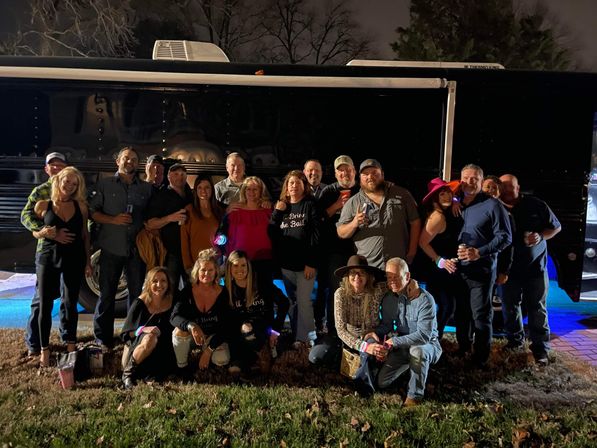 About 20 friends posing outside a black party/tour bus at night on a grassy lawn, smiling and holding drinks in casual and festive outfits.
