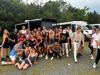 Large group of friends in summer outfits posing and smiling in front of parked party buses on a gravel lot with trees in the background — festival shuttle scene.