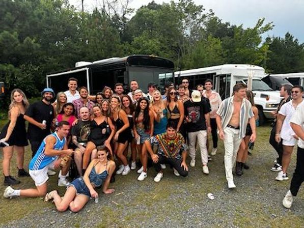 Large group of friends in summer outfits posing and smiling in front of parked party buses on a gravel lot with trees in the background — festival shuttle scene.