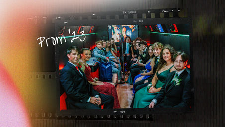 Group of teens in formal dresses and suits seated along both sides of a party-bus interior, smiling and posing for a prom photo under colorful LED lighting and a center pole, with handwritten "Prom '23" overlay.