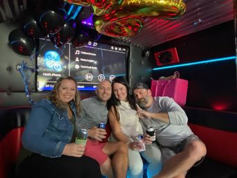 Four friends smiling and holding drinks in a party-bus karaoke lounge, seated on black-and-red leather bench under gold balloons and blue LED lights with a large music screen and pink gift bag nearby.