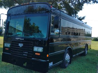 Sleek black passenger bus parked on a grassy field beneath trees, front three-quarter view with reflective side panels and chrome wheel