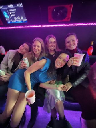 Five friends smiling and holding drinks inside a neon-lit party bus lounge with purple LED lights, nightlife vibe
