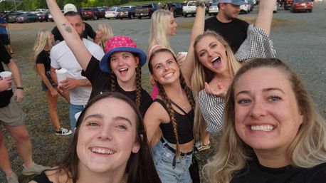 Five friends taking a cheerful selfie at an outdoor music festival parking lot, smiling and posing with rows of parked cars in the background.