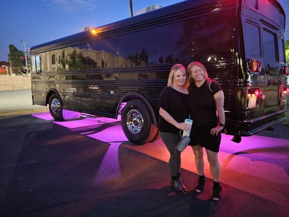 Two smiling women posing in front of a black party bus with pink-purple underglow lights in a parking lot at dusk, one holding a cup — night out vibe.