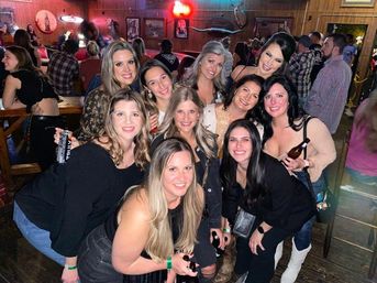 Group of smiling women posing together at a crowded rustic country bar with wood-paneled walls, neon lights and a mounted deer head in the background, several holding beer bottles and cans.