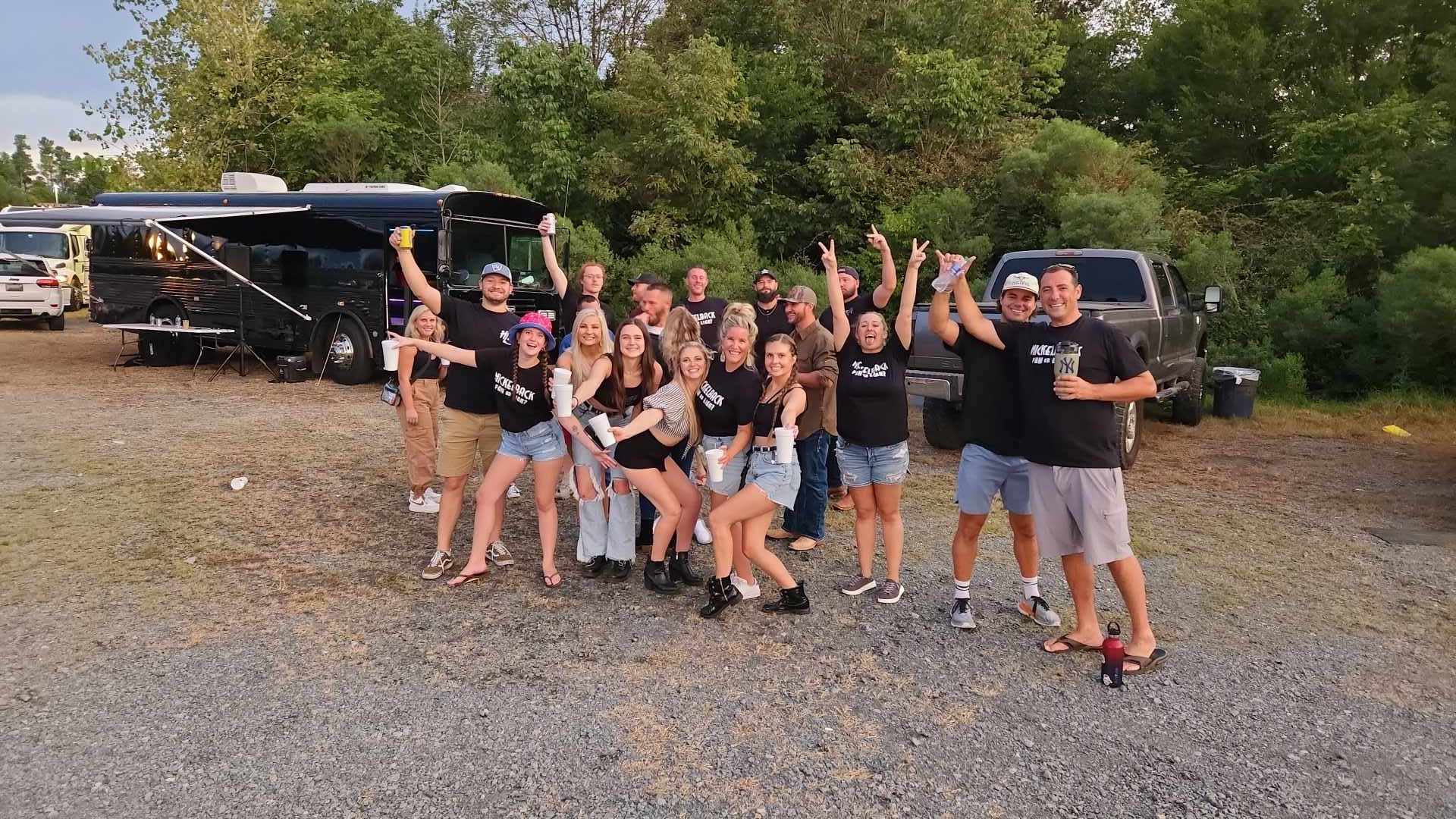 Cheerful group of friends posing for a group photo while tailgating near a black tour bus and pickup truck in a wooded campground parking area