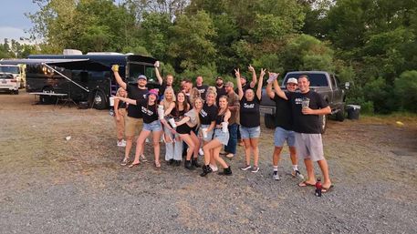 Cheerful group of friends at an outdoor tailgate/campground gathering by a black tour bus and pickup truck, posing with drinks and celebratory gestures amid trees and gravel