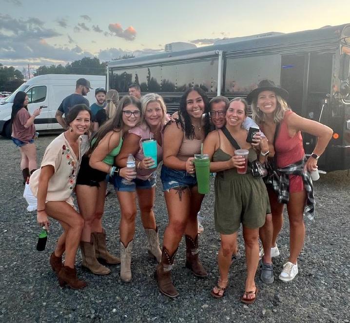 Group of eight friends smiling and posing at an outdoor music festival tailgate in a gravel lot at sunset, wearing summer outfits and cowboy boots and holding colorful drinks in front of a black tour bus.