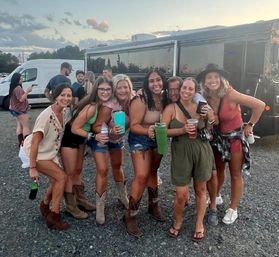 Group of seven women smiling and holding drinks in a gravel concert parking lot at sunset, wearing summer outfits and cowboy boots beside a tour bus for an outdoor tailgate.