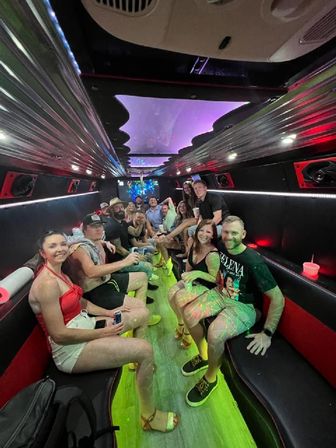 Group of friends partying inside a neon-lit party bus interior, seated on long benches with drinks, LED ceiling panels and green floor lighting for nightlife vibes.