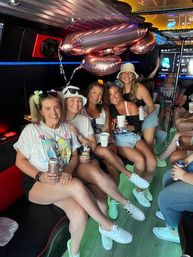 Smiling group of friends in summer outfits sitting inside a party bus, holding drinks with metallic balloons overhead and casual sneakers